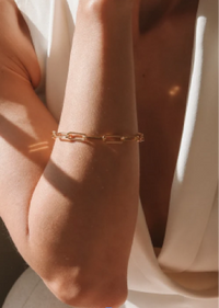 A close-up of a delicate gold chain bracelet worn on a person's wrist, styled with a white draped garment under warm, direct sunlight.