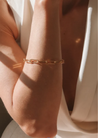 A close-up of a delicate gold chain bracelet worn on a person's wrist, styled with a white draped garment under warm, direct sunlight.