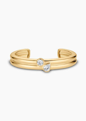 A gold-toned open cuff ring featuring two small clear gemstones, one round and one pear-cut, set against a plain white background.
