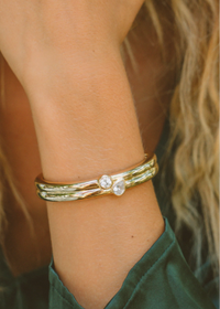 A close-up of a person's wrist wearing a gold-toned multi-band bracelet with two small clear round stones at the center, against a warm blurred background.