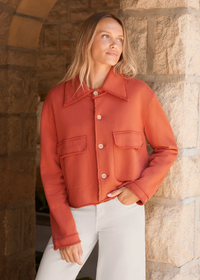A woman wearing a cropped, rust-orange button-up jacket with a pointed collar and flap chest pockets, paired with white pants, standing in front of a rustic stone archway.