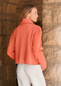 A woman wearing a cropped, long-sleeve coral-pink fleece jacket and white pants, standing in front of a rustic stone wall.