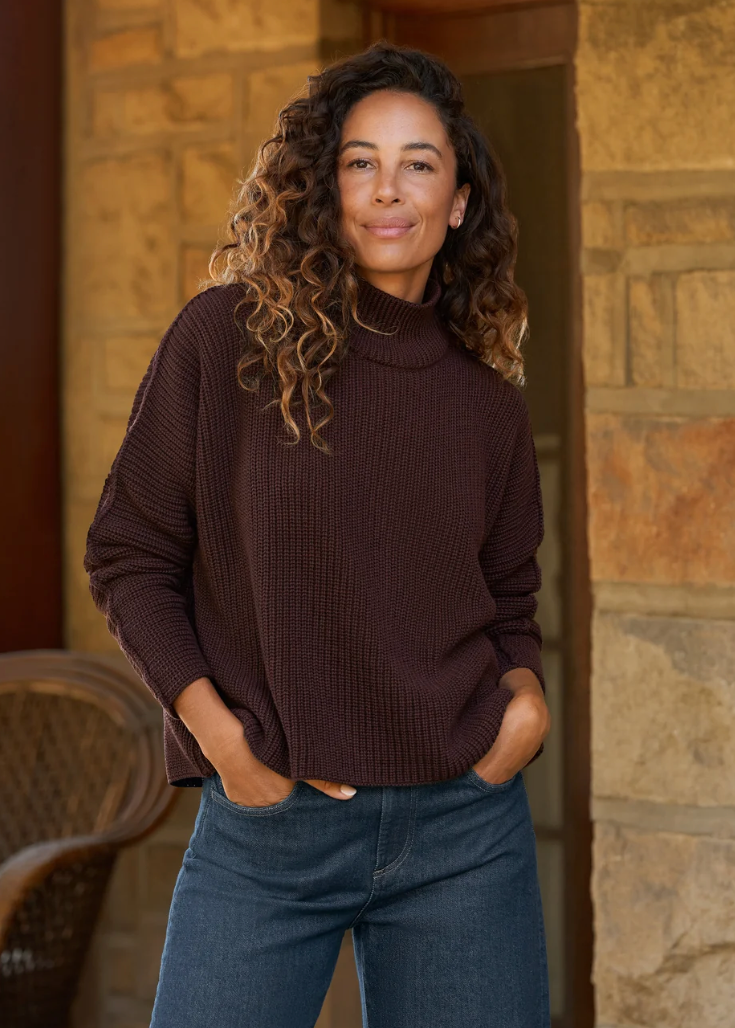 A woman with curly brown hair wearing a dark brown ribbed turtleneck sweater and blue jeans, standing in front of a stone wall doorway.