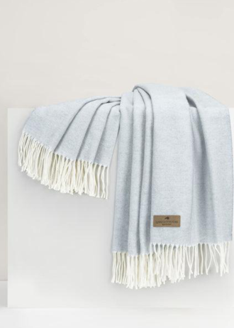 A light blue woven throw blanket with white fringe, draped over a white display block against a neutral off-white background. A small brown leather brand tag is visible near the bottom right.