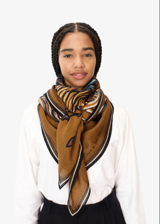 A woman wearing a white blouse and dark skirt, styled with a brown silk scarf featuring black, gold, and blue patterns, set against a plain white background.