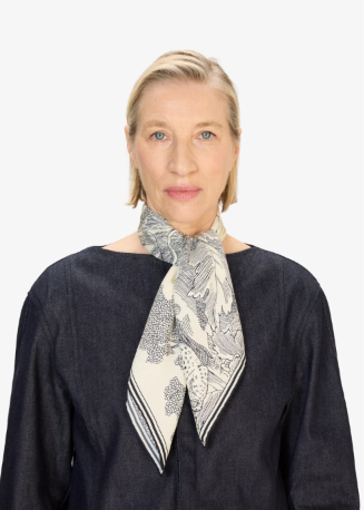 A woman with short blonde hair wearing a dark navy blue top and a cream-colored silk scarf with a black botanical pattern, photographed against a solid white background.