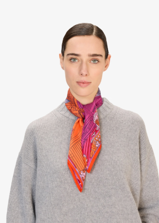 A woman wearing a gray knit sweater and a bright patterned silk scarf in orange, pink, and purple, photographed against a clean white background.