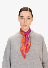 A woman wearing a gray knit sweater and a bright patterned silk scarf in orange, pink, and purple, photographed against a clean white background.