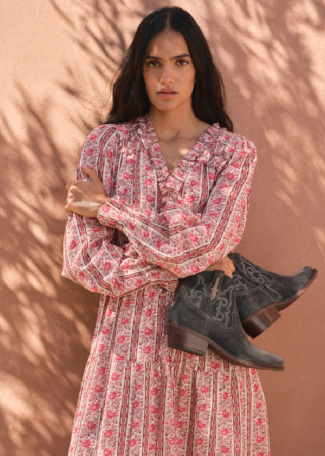 A woman stands against a warm terracotta wall with leaf shadows, wearing a long-sleeve floral midi dress and holding a pair of dark gray western-style boots.
