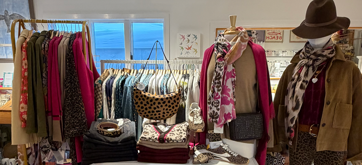 A boutique clothing display featuring a mannequin dressed in a brown hat, brown jacket, burgundy top, leopard-print scarf, and patterned scarf, alongside mannequins and racks of colorful apparel in a retail showroom.