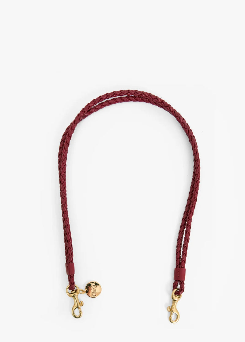 A braided leather shoulder strap in burgundy with gold-tone hardware, including a small decorative charm and two snap hooks, shown on a plain white background.
