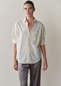 A model wearing a cream-colored, long-sleeve button-down shirt with rolled-up sleeves and grey trousers, standing against a plain beige background.