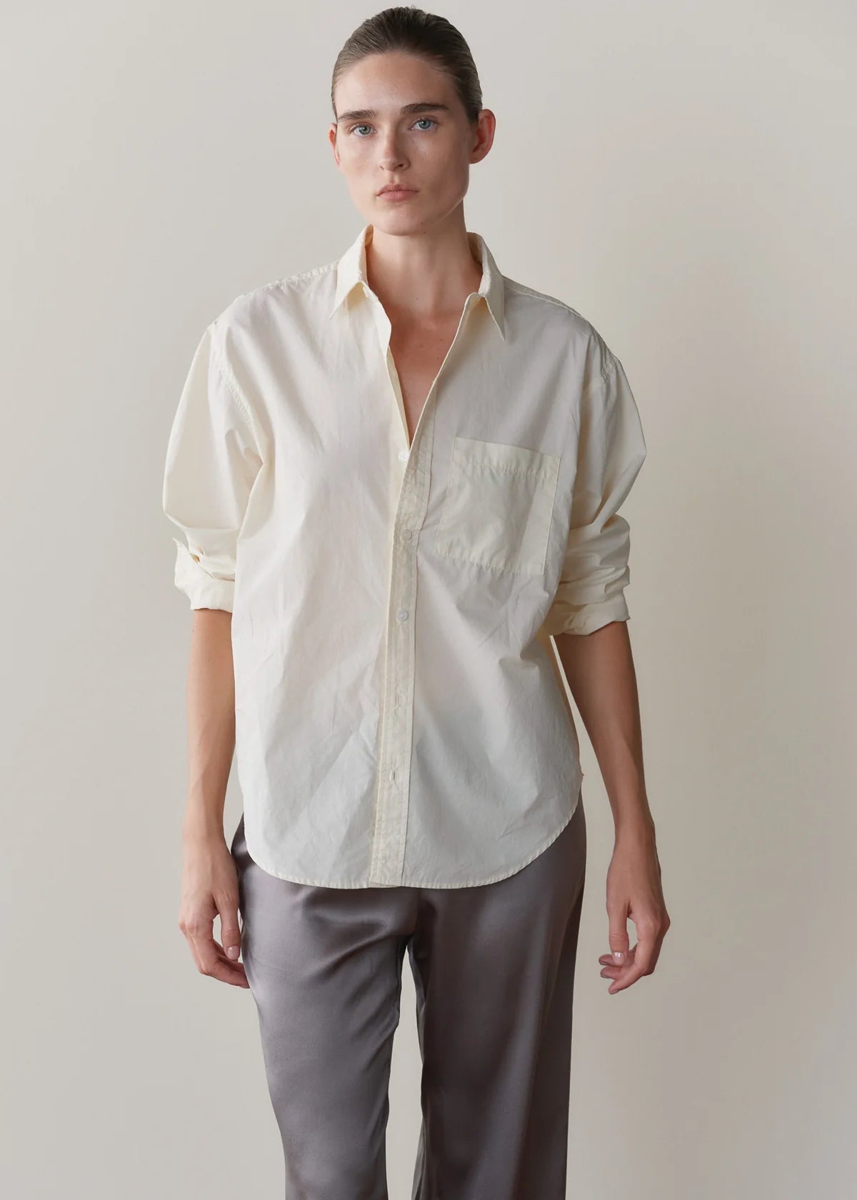 A model wearing a cream-colored, long-sleeve button-down shirt with rolled-up sleeves and grey trousers, standing against a plain beige background.