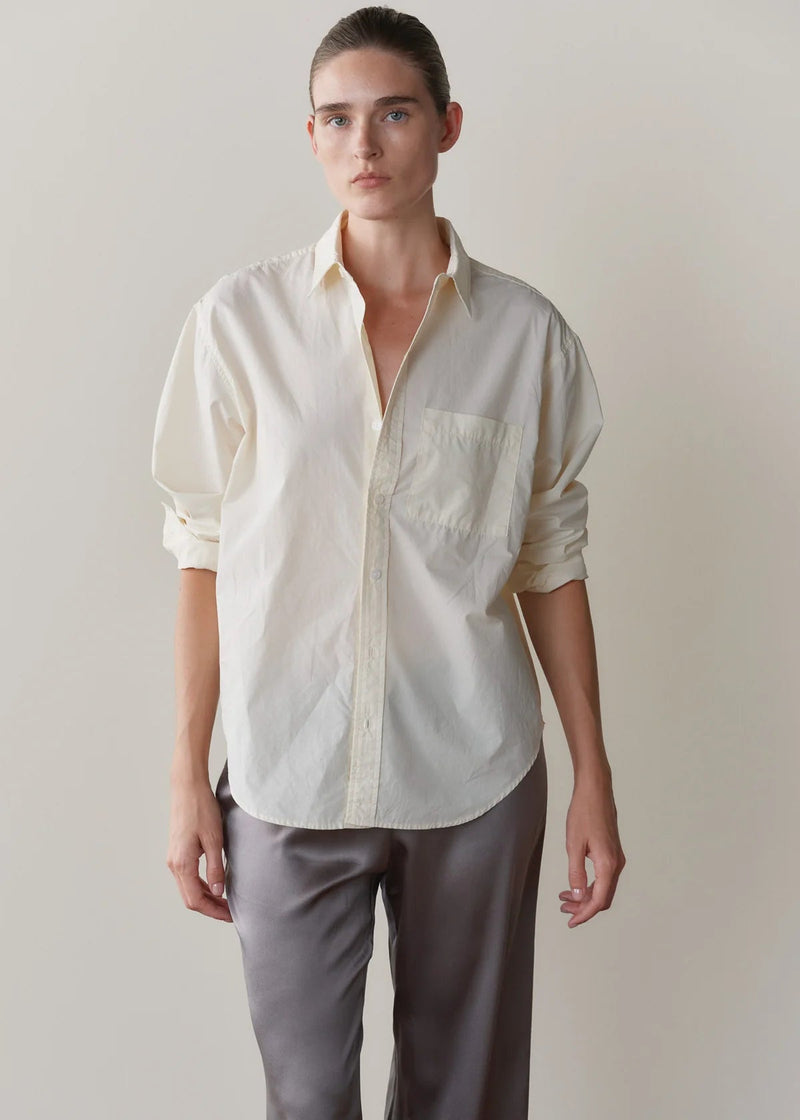 A model wearing a cream-colored, long-sleeve button-down shirt with rolled-up sleeves and grey trousers, standing against a plain beige background.