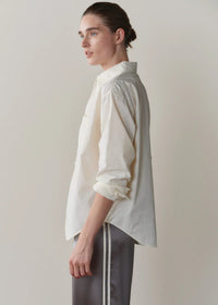 A model shown in profile wearing a cream-colored, long-sleeve button-up shirt with rolled-up sleeves and dark gray pants with white side stripes, set against a plain light gray studio background.