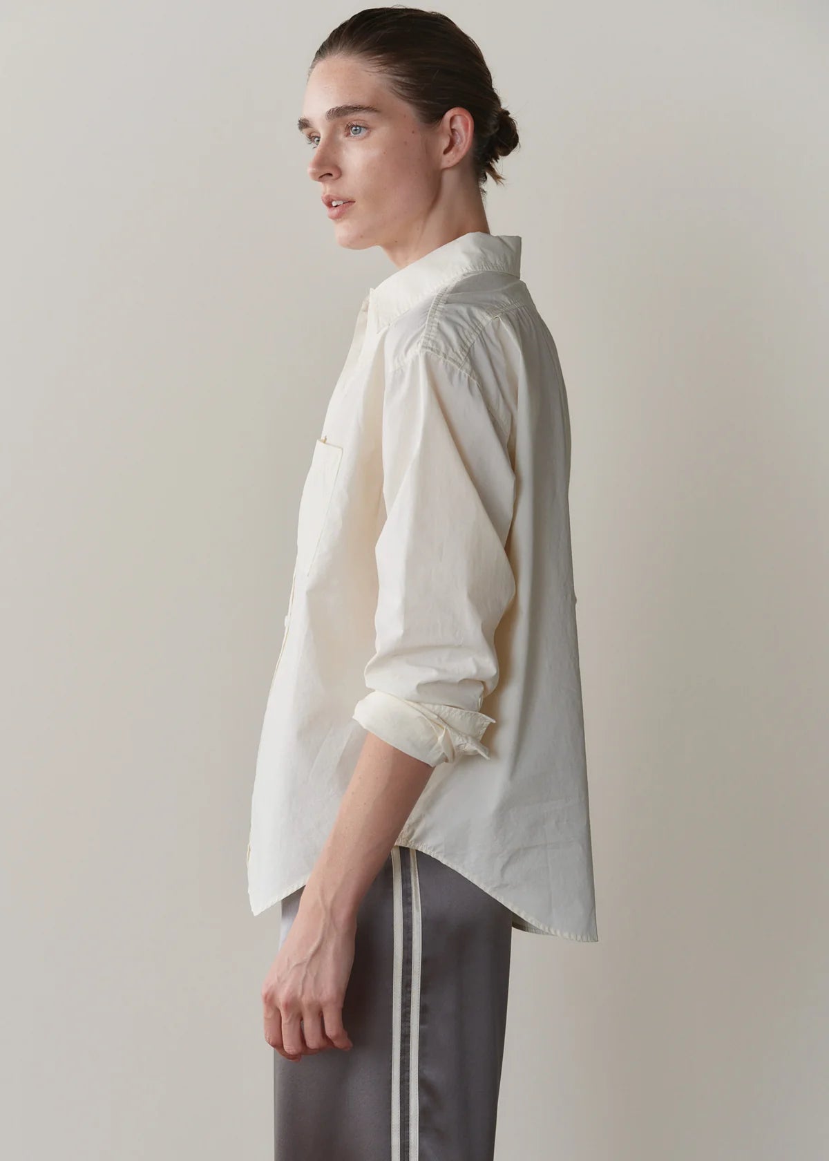 A model shown in profile wearing a cream-colored, long-sleeve button-up shirt with rolled-up sleeves and dark gray pants with white side stripes, set against a plain light gray studio background.