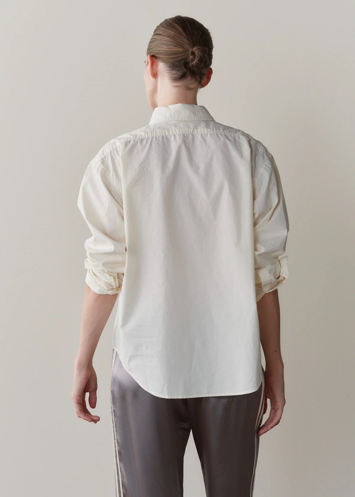 A woman shown from the back wearing a cream-colored, long-sleeve button-up shirt with gathered cuffs and dark gray pants, set against a plain light beige studio background.