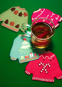 Four Christmas sweater-shaped fabric coasters in green, pink, blue, and red, arranged on a solid green background with a glass mug filled with iced tea on top.