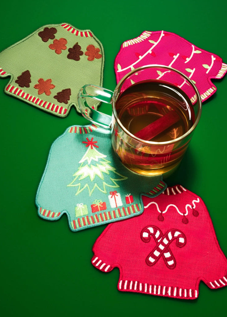 Four Christmas sweater-shaped fabric coasters in green, pink, blue, and red, arranged on a solid green background with a glass mug filled with iced tea on top.
