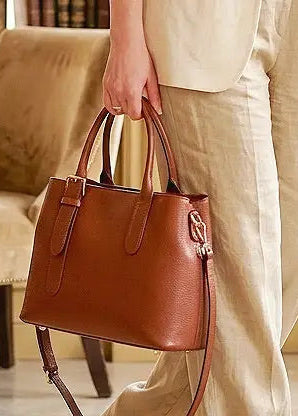 A person in a cream-colored outfit holds a brown pebbled leather handbag with double top handles and a shoulder strap in an indoor setting.
