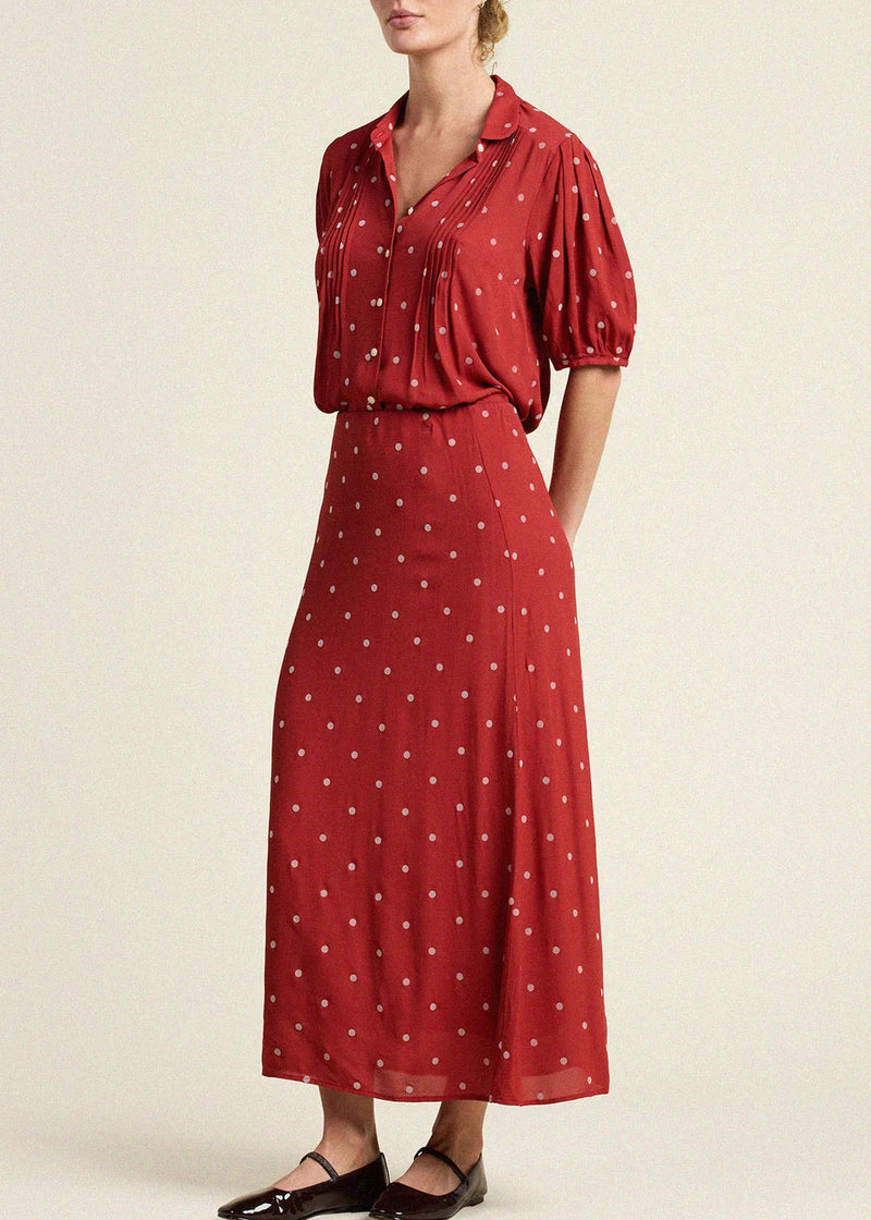 A woman models a red midi dress with white polka dots, featuring a button-down collar, short puff sleeves, and a flowing skirt, paired with black flat shoes against a light beige background.