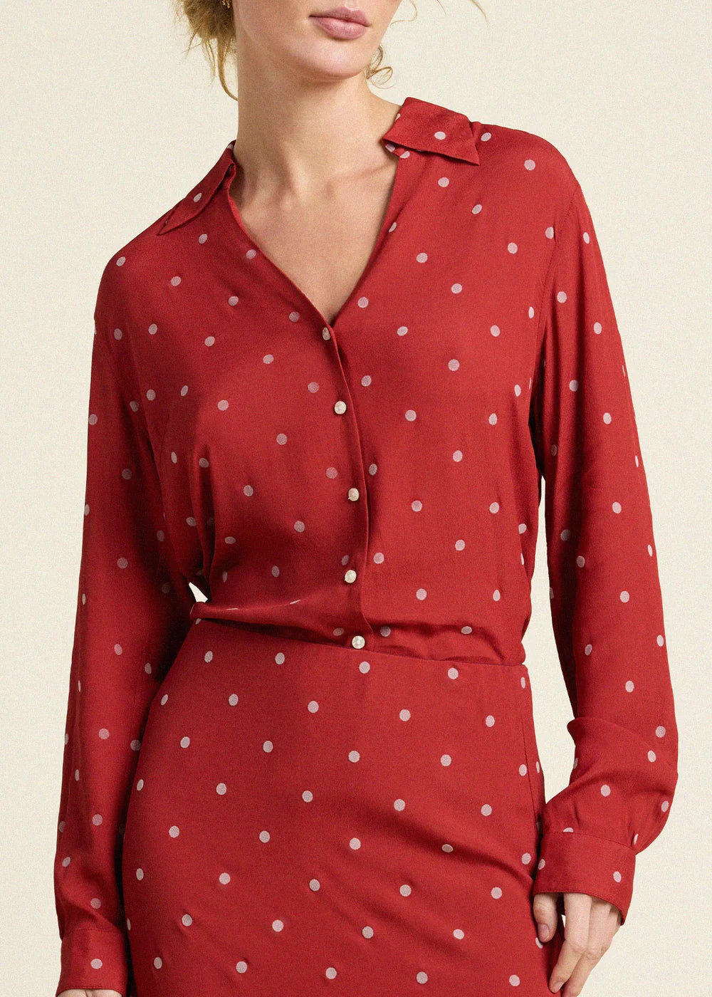 A woman wearing a red long-sleeve collared blouse and matching skirt set, both with a small white polka dot pattern, against a plain light beige background.