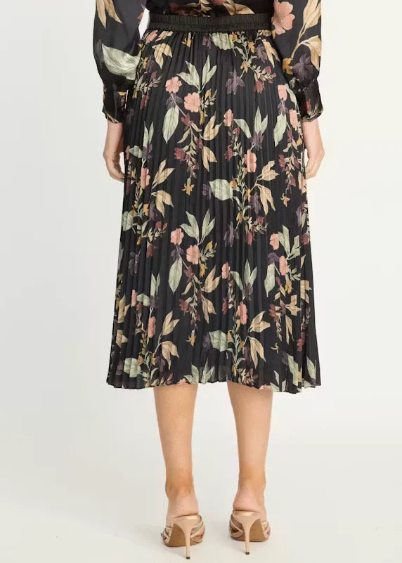 A woman wearing a black pleated midi skirt with a floral print in muted pink, green, and beige tones, paired with beige heeled sandals against a plain light background.