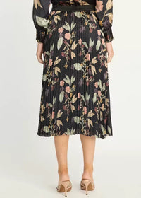 A woman wearing a black pleated midi skirt with a floral print in muted pink, green, and beige tones, paired with beige heeled sandals against a plain light background.