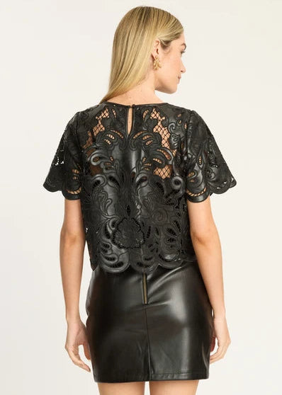 A woman shown from the back wearing a black lace short-sleeve top with a floral lace overlay and a black faux-leather mini skirt, against a plain light background.