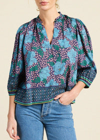 A woman wearing a short-sleeve blouse with a blue, green, and purple floral pattern, paired with blue jeans, against a light beige background.