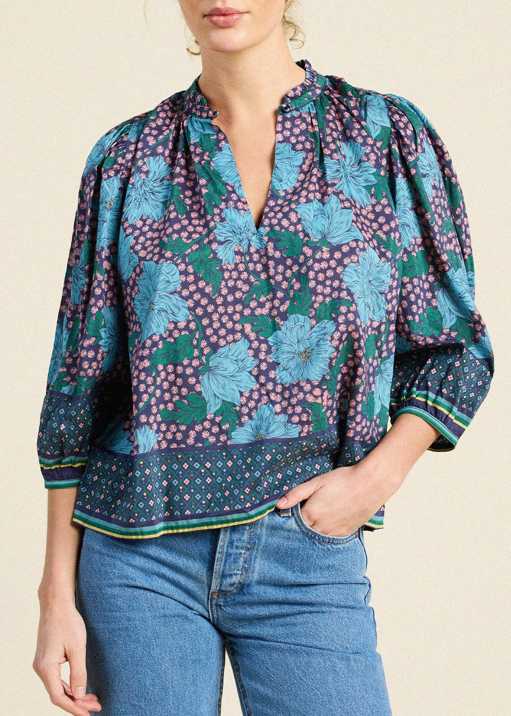 A woman wearing a short-sleeve blouse with a blue, green, and purple floral pattern, paired with blue jeans, against a light beige background.