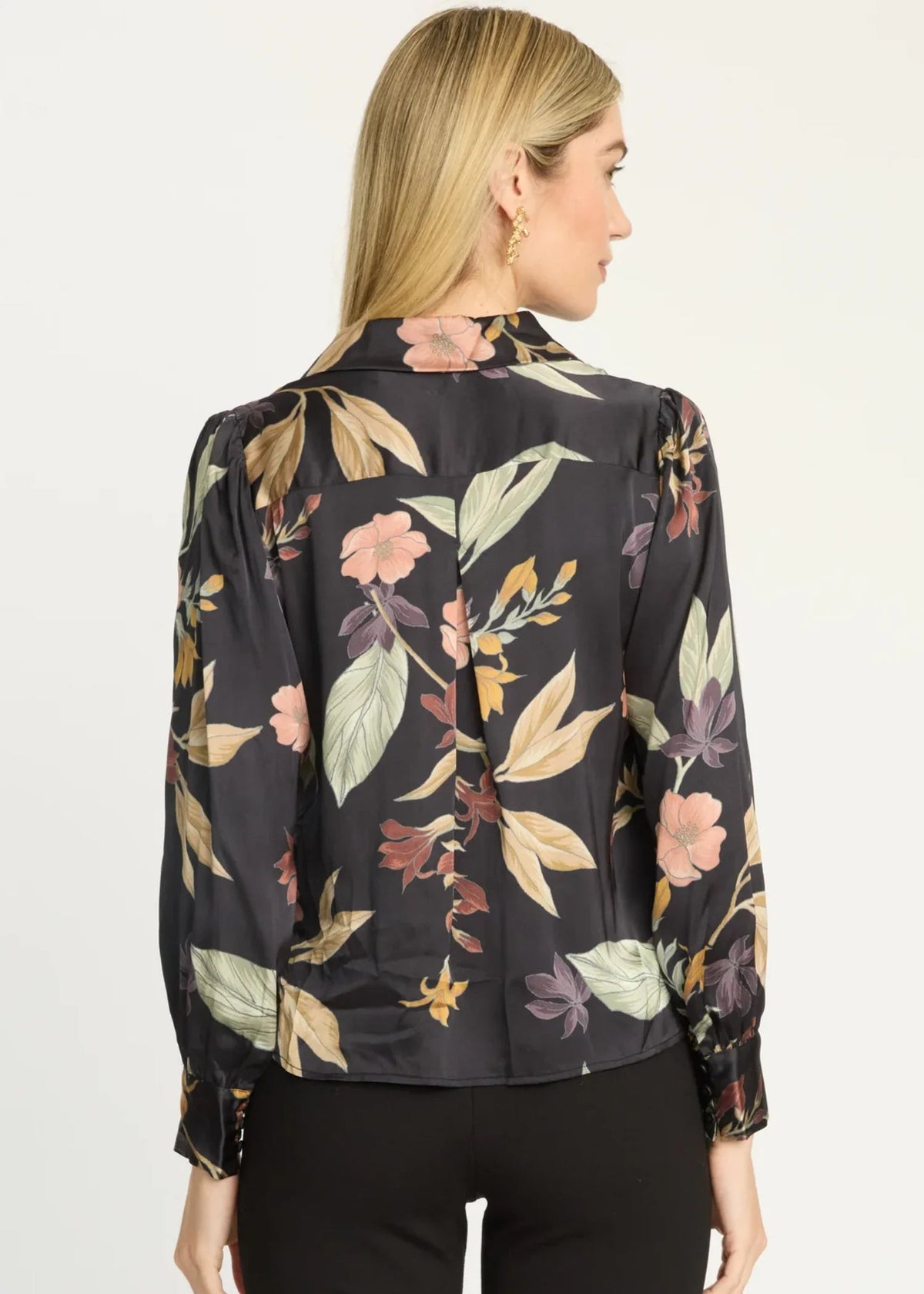 A woman shown from the back wearing a black long-sleeve blouse with a large floral print in muted pink, beige, and olive green, paired with black pants against a plain white studio background.