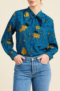 A woman wearing a teal blue long-sleeve blouse with a yellow floral print and a bow collar, paired with blue jeans, against a light beige background.