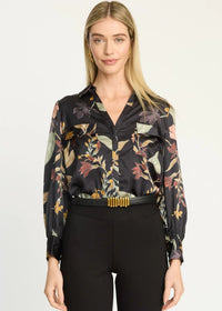 A woman modeling a black long-sleeve button-up blouse with a multicolor floral and botanical print, paired with black pants and a black belt with gold accents, against a plain light background.